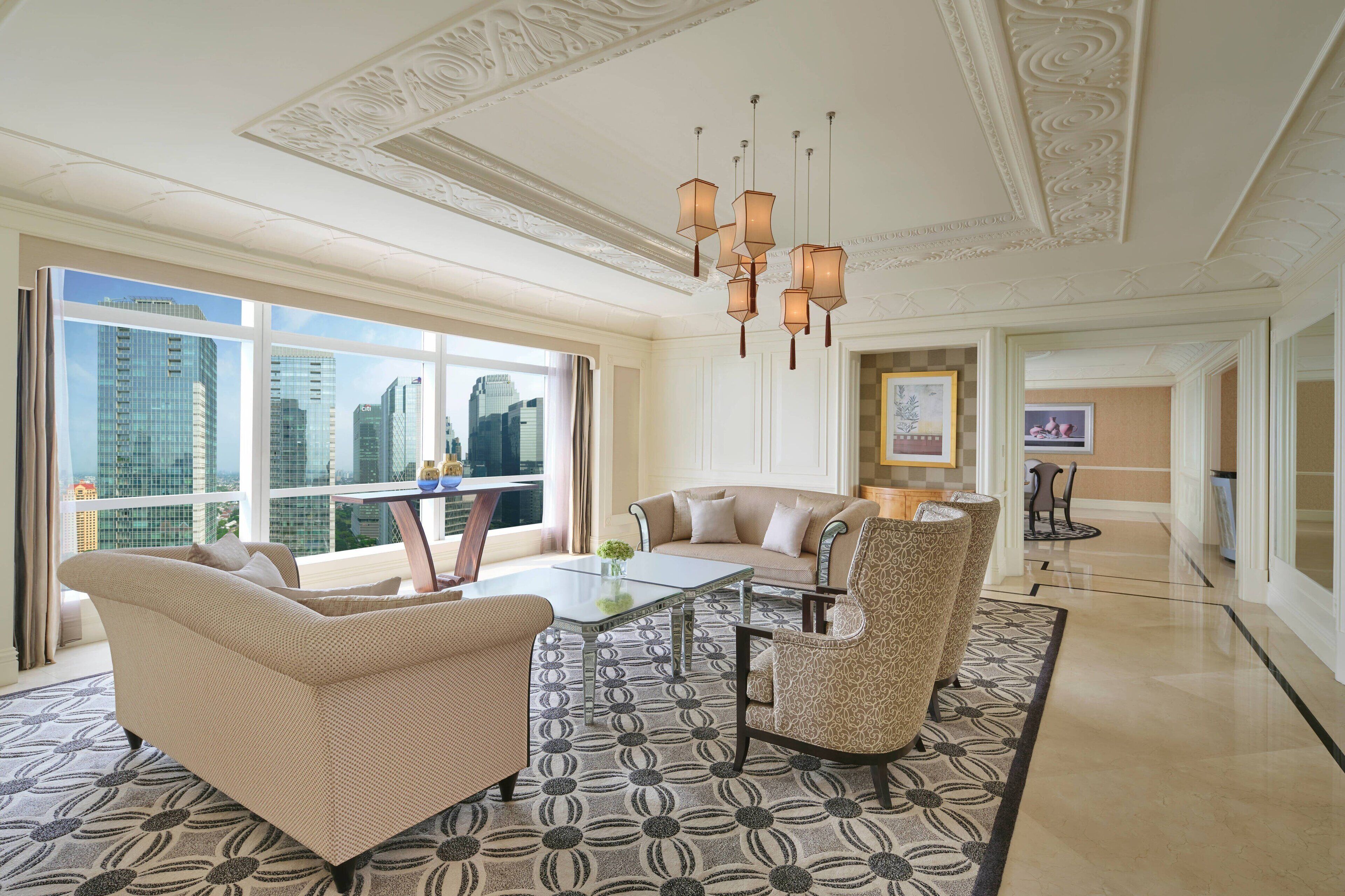 A spacious living room with a high ceiling, a large window offering a city view, a patterned rug, and a modern chandelier.
