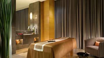 A modern spa room with a massage table, a sofa, and a large window with curtains.