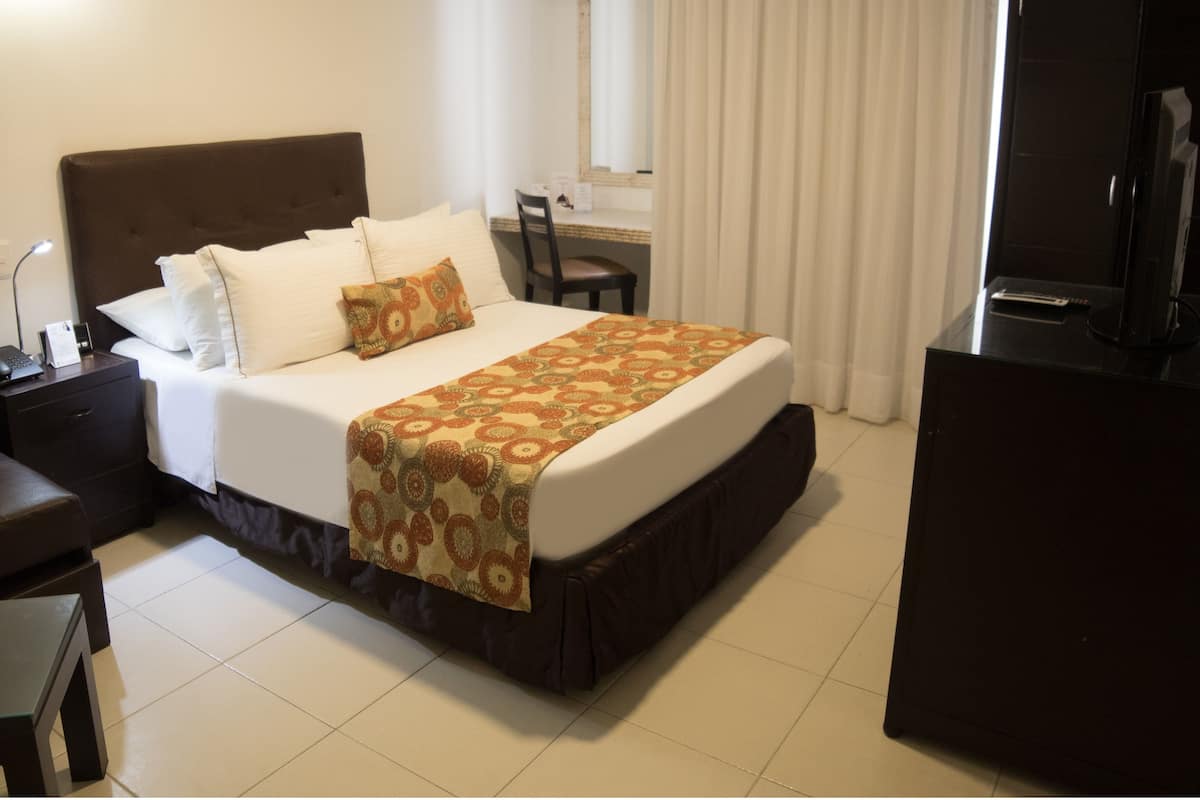 A hotel room with a large bed, a desk, a chair, an air conditioning unit, and a television.