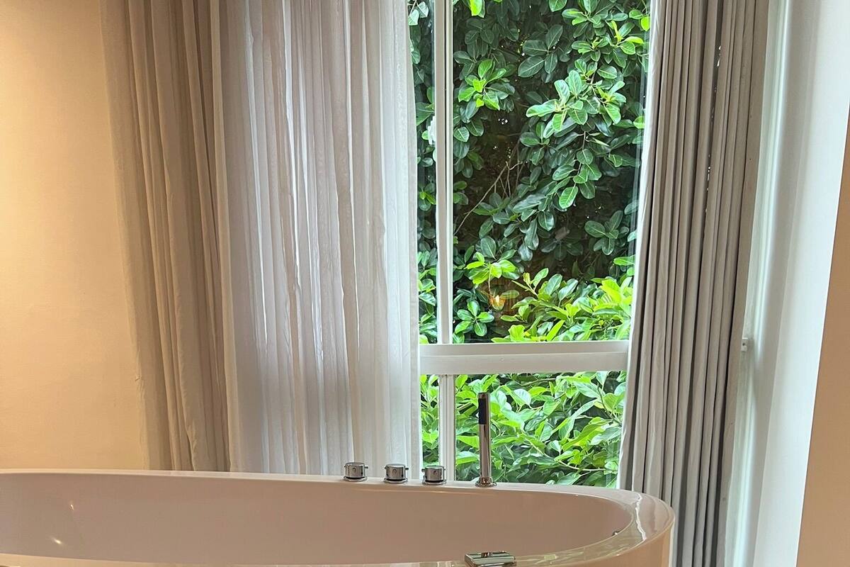 A modern bathroom with a freestanding bathtub, a stone accent wall, and a view of greenery through large windows.