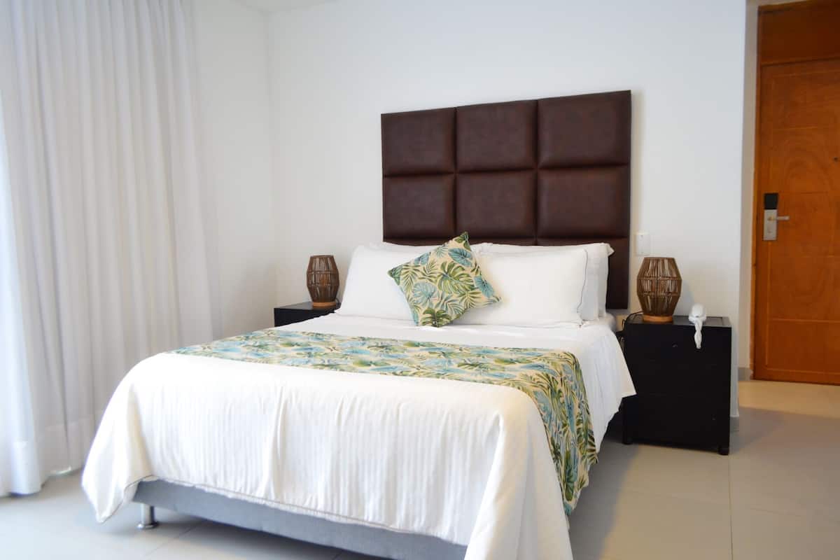 A neatly made bed with a white duvet, a decorative pillow, and a patterned bedspread. There are two bedside tables with lamps and a wooden door in the background.