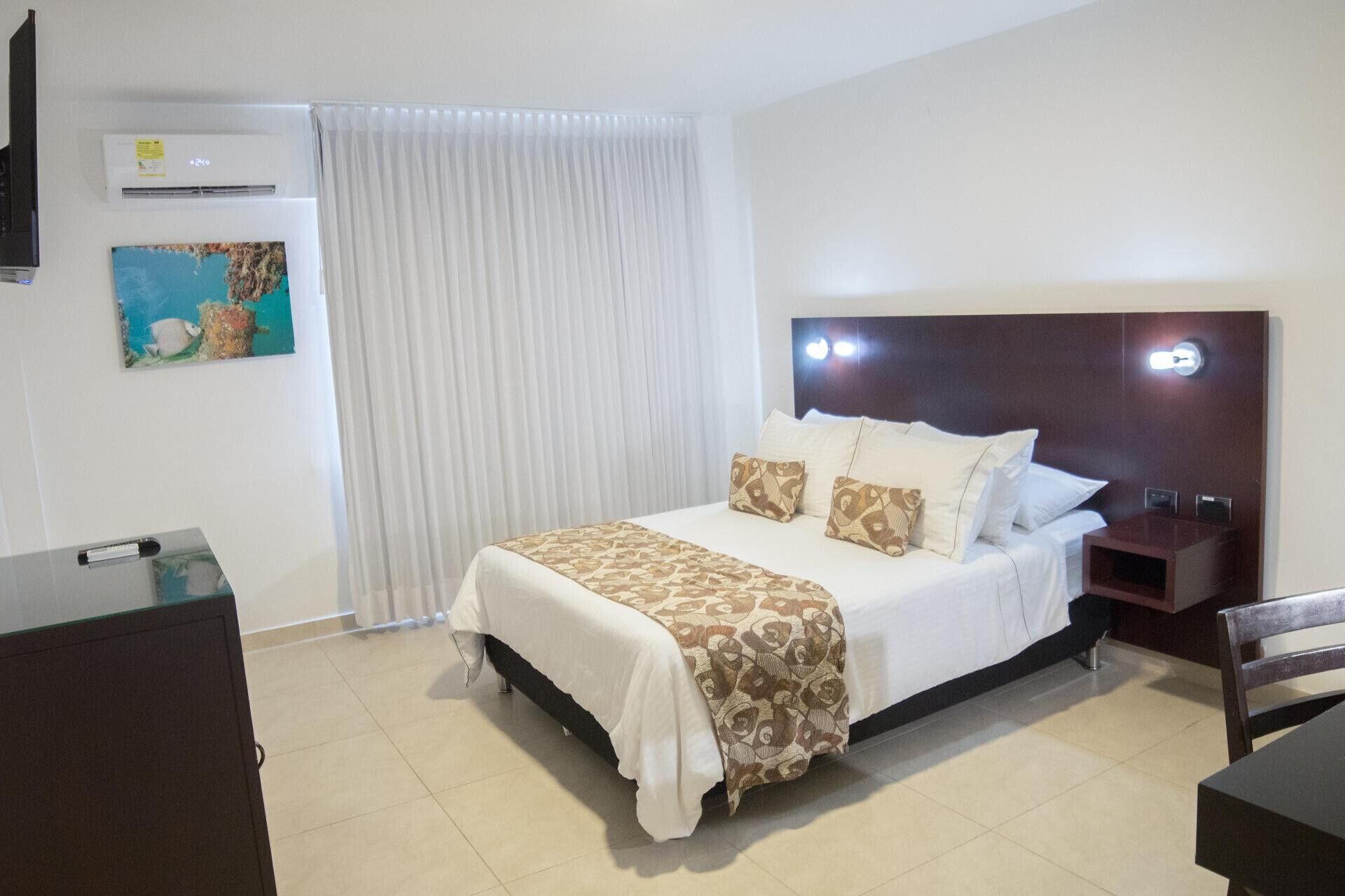 A hotel room with a bed, a nightstand, a desk, an air conditioner, and a painting on the wall.