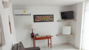 Un salon avec un canapé marron, une petite table avec une lampe, un téléviseur posé sur un meuble et un réfrigérateur.