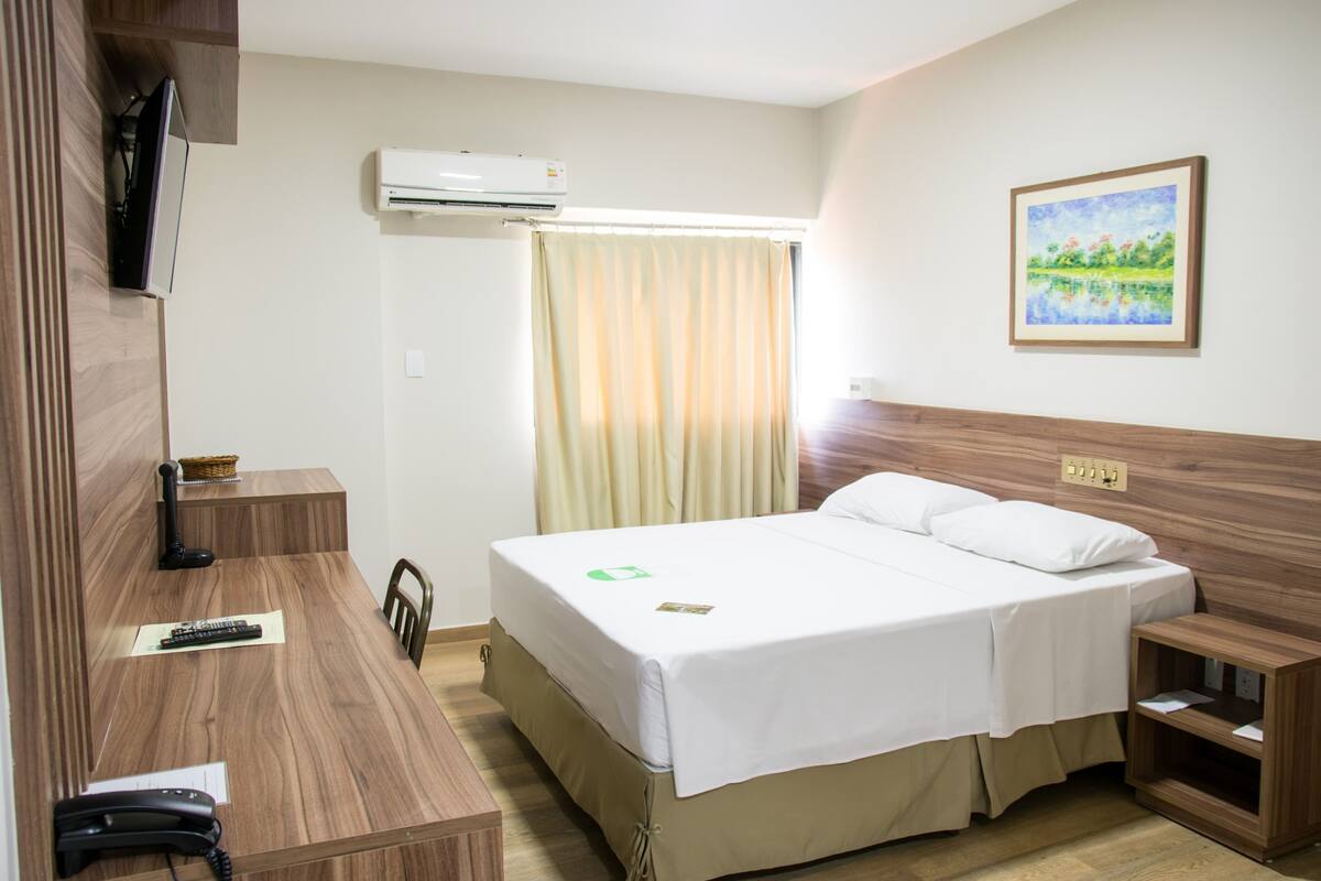 A hotel room with a bed, bedside table, a desk with a telephone, a chair, an air conditioner, a window with curtains, and a framed picture on the wall.