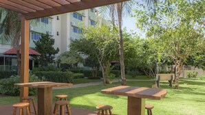 An outdoor patio area with wooden tables and stools, surrounded by greenery and a building in the background. - Hotel Vale Verde (Campo Grande)