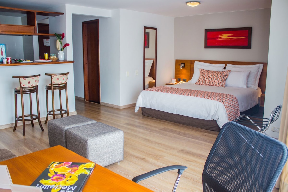 A hotel room with a bed, a desk, two chairs, a small table, and a mirror.