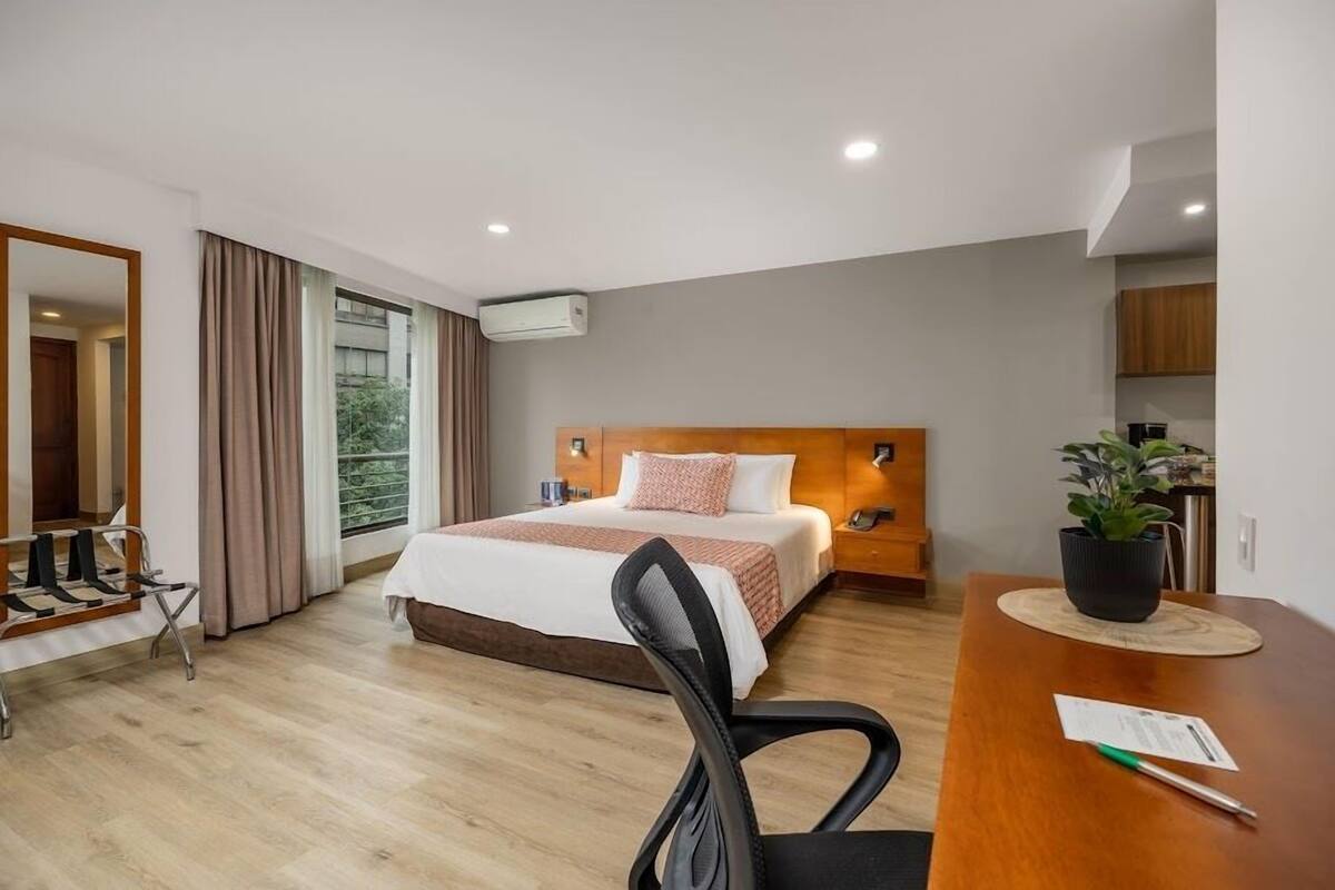 Ein modernes Hotelzimmer mit einem großen Bett, einem Holz-Kopfteil, einem Schreibtisch mit Stuhl, einer Topfpflanze und Blick ins Freie.