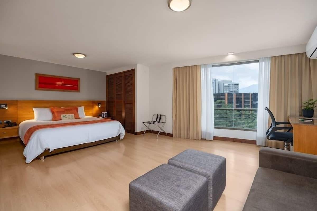 Ein modernes Hotelzimmer mit einem großen Bett, einem Schreibtisch, einem Sessel und Blick auf eine Stadtlandschaft.