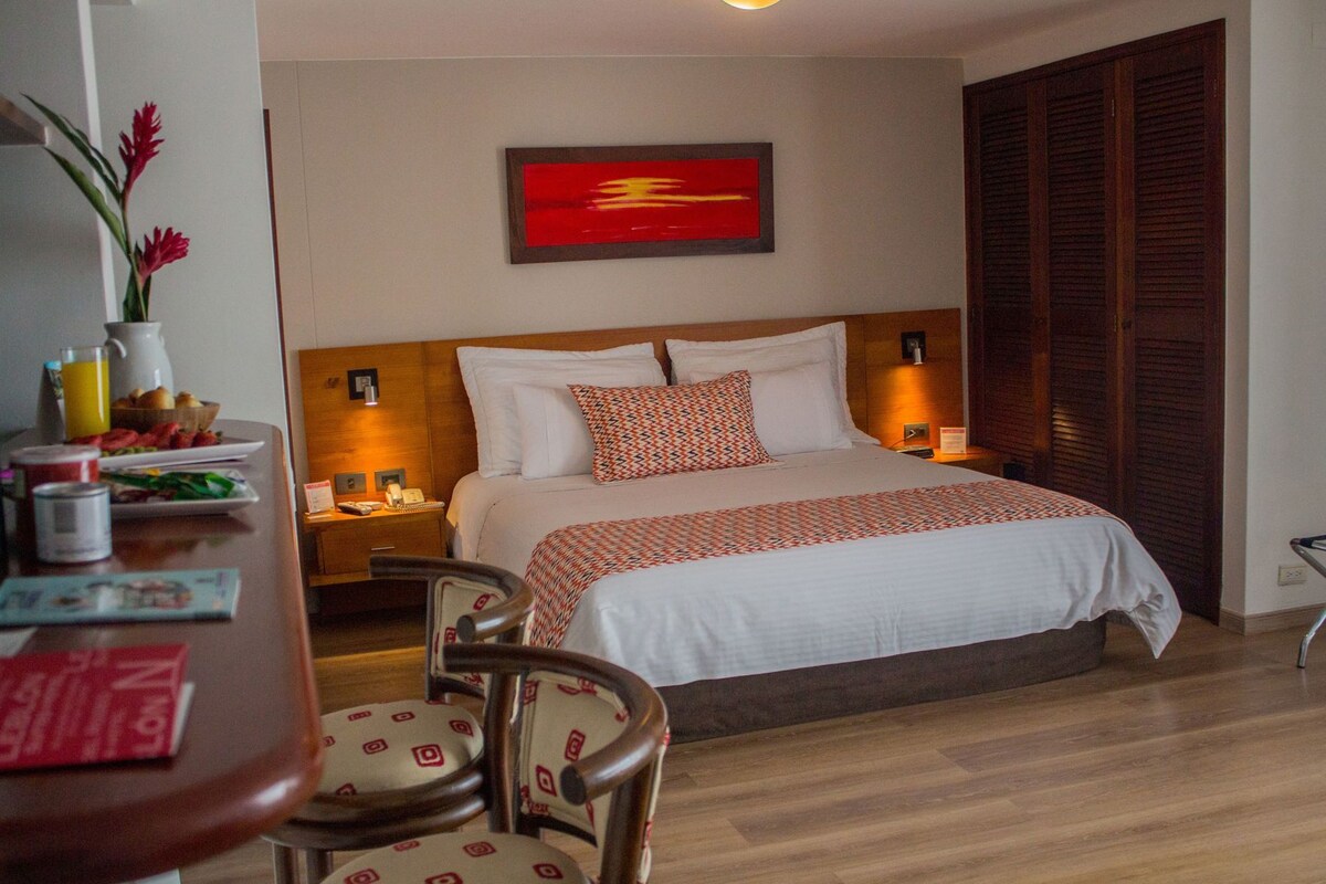 A hotel room with a bed, bedside tables, a desk with chairs, a wall-mounted painting, and a wooden wardrobe.