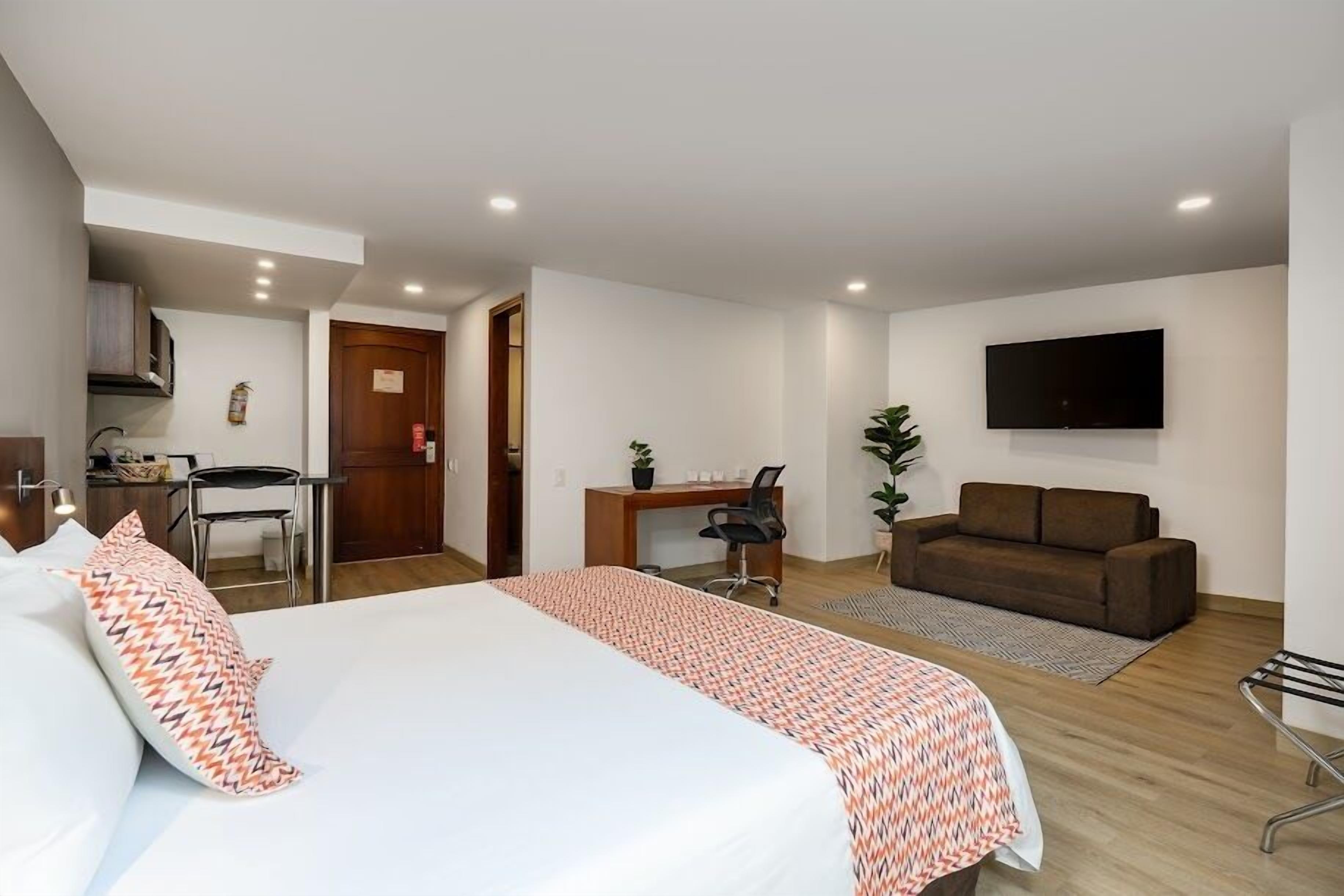 Habitación de hotel con cama, escritorio, sillón, sofá, televisión y una pequeña kitchenette.