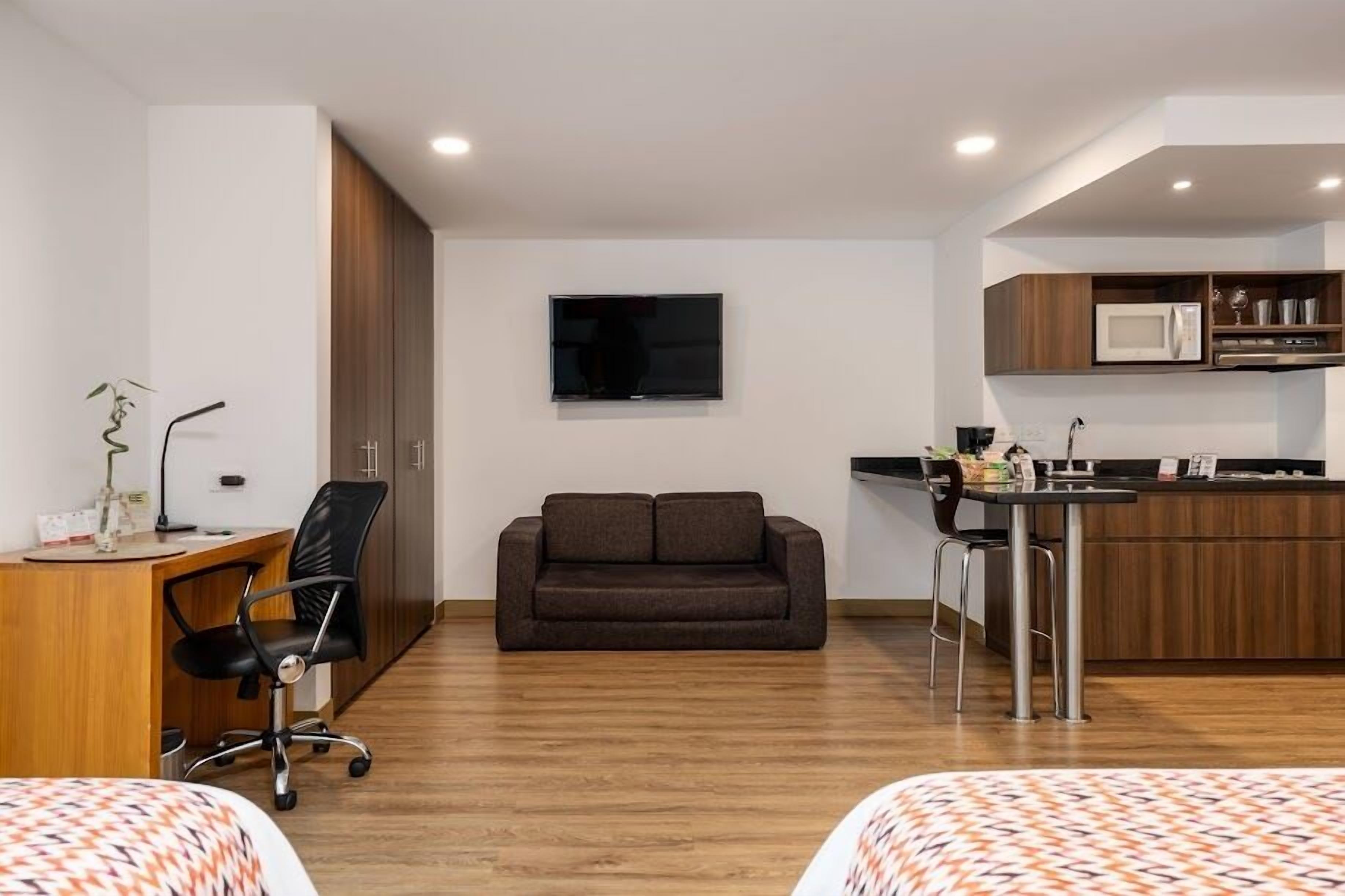 A modern hotel room with a wooden desk, a sofa, a TV, a kitchenette, and a bed with patterned bedding.