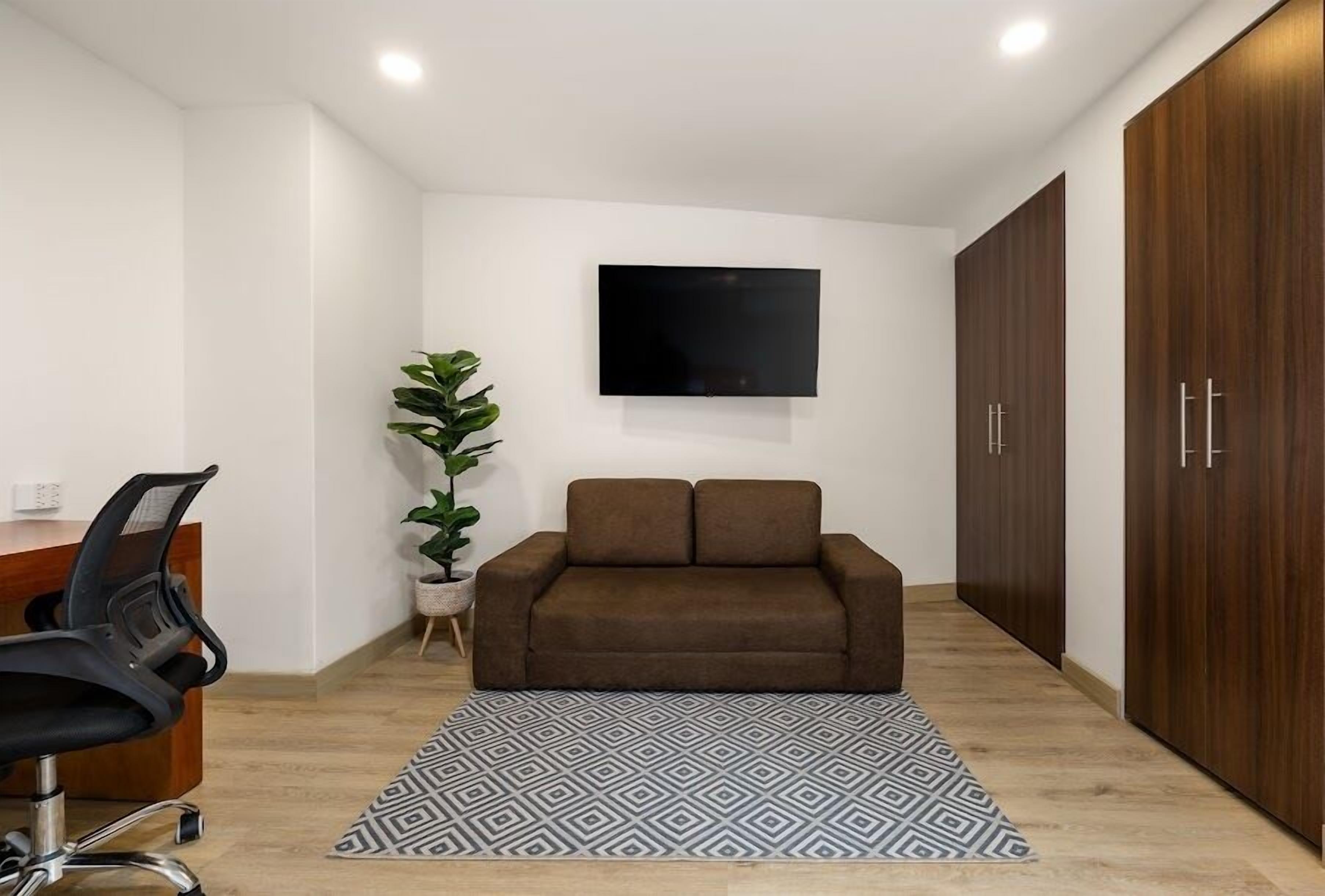 A modern office space with a brown sofa, a desk with a chair, a potted plant, and a patterned rug.