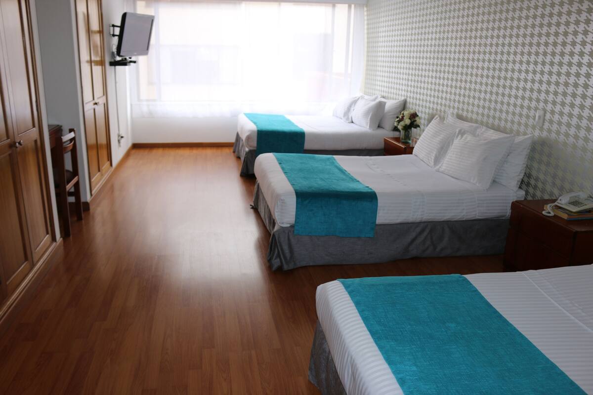 Habitación de hotel con dos camas, piso de madera, un televisor montado en la pared y un papel tapiz estampado.