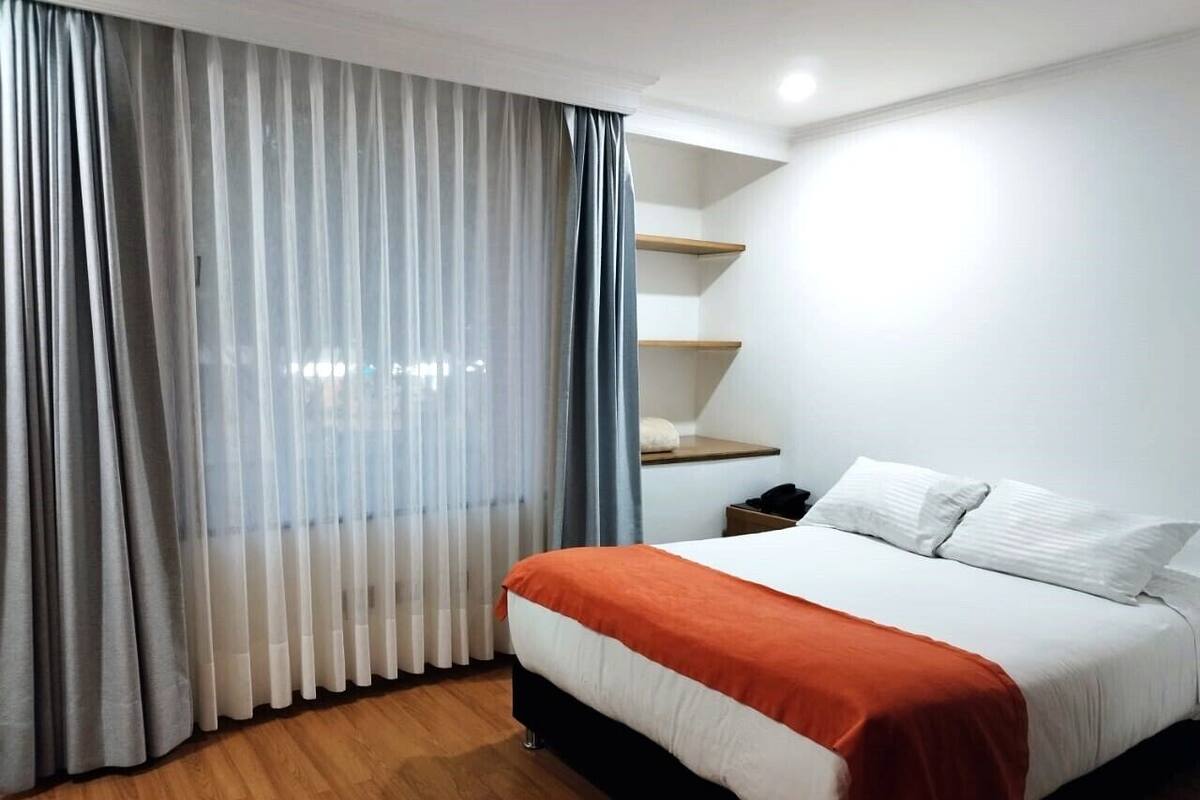 Una habitación de hotel con cama, suelo de madera, mesita de noche y ventana con cortinas translúcidas.