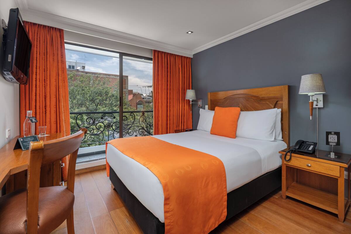 Habitación de hotel con una cama grande, cortinas anaranjadas, un escritorio de madera y vistas al exterior.