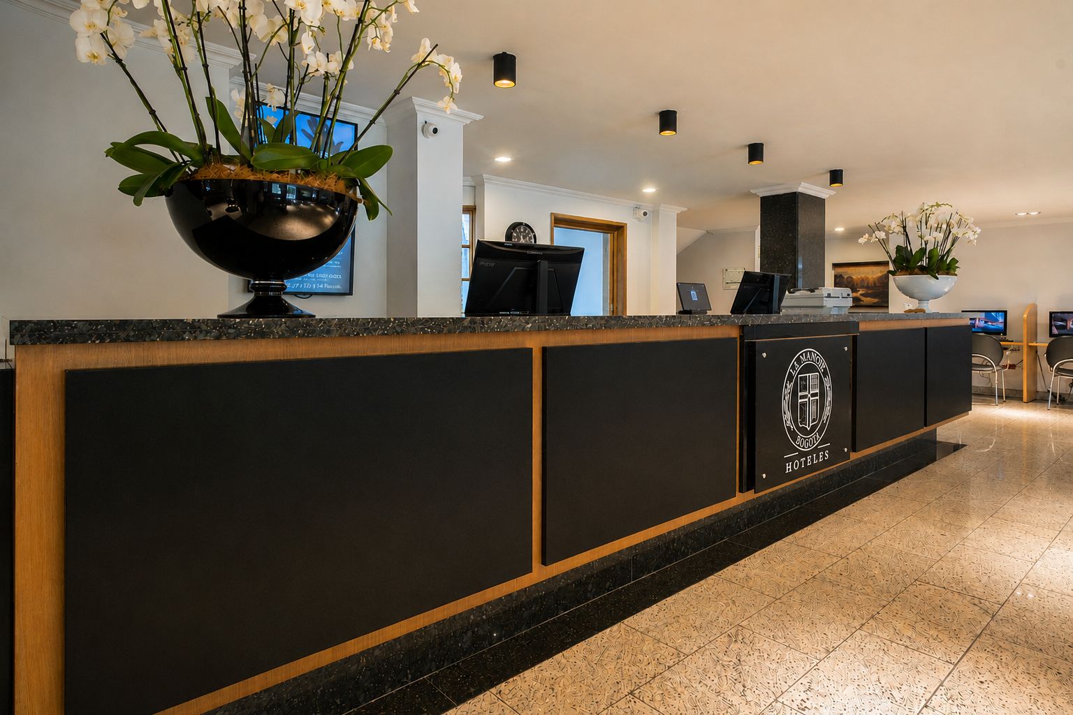 Una recepción de hotel con un logotipo, una maceta negra grande con flores blancas y una pantalla digital.