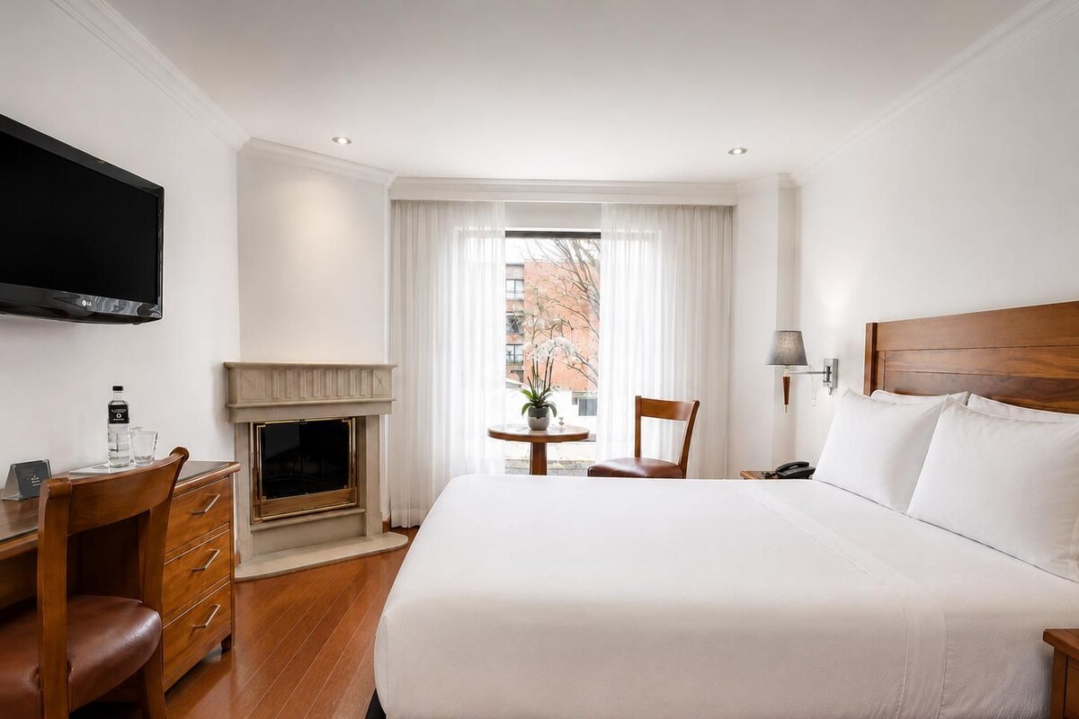 Una habitación de hotel con una cama grande, un escritorio con silla, televisión, chimenea, ventana con cortinas y una lámpara.