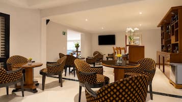 A hotel lobby with a seating area featuring patterned chairs and tables, a flat-screen TV mounted on the wall, and a wooden cabinet with various items.