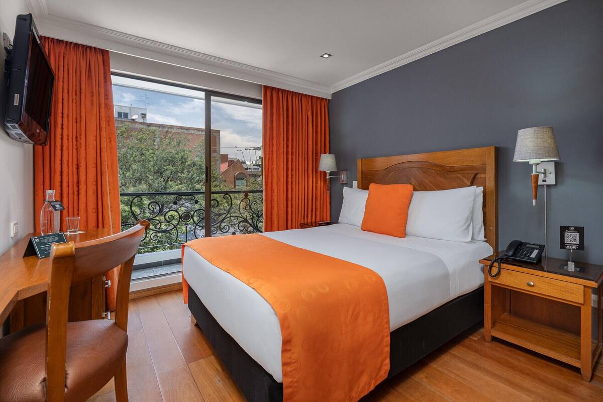 Habitación de hotel con cama, escritorio, silla, mesita de noche, lámpara y vistas al exterior.
