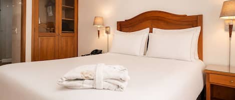 A hotel room with a large bed, wooden headboard, two bedside lamps, a wooden nightstand, and a glass door leading to a bathroom.