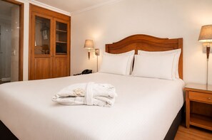 Une chambre d’hôtel avec un grand lit, une tête de lit en bois, deux lampes de chevet, une table de chevet en bois et une porte en verre donnant sur la salle de bain.