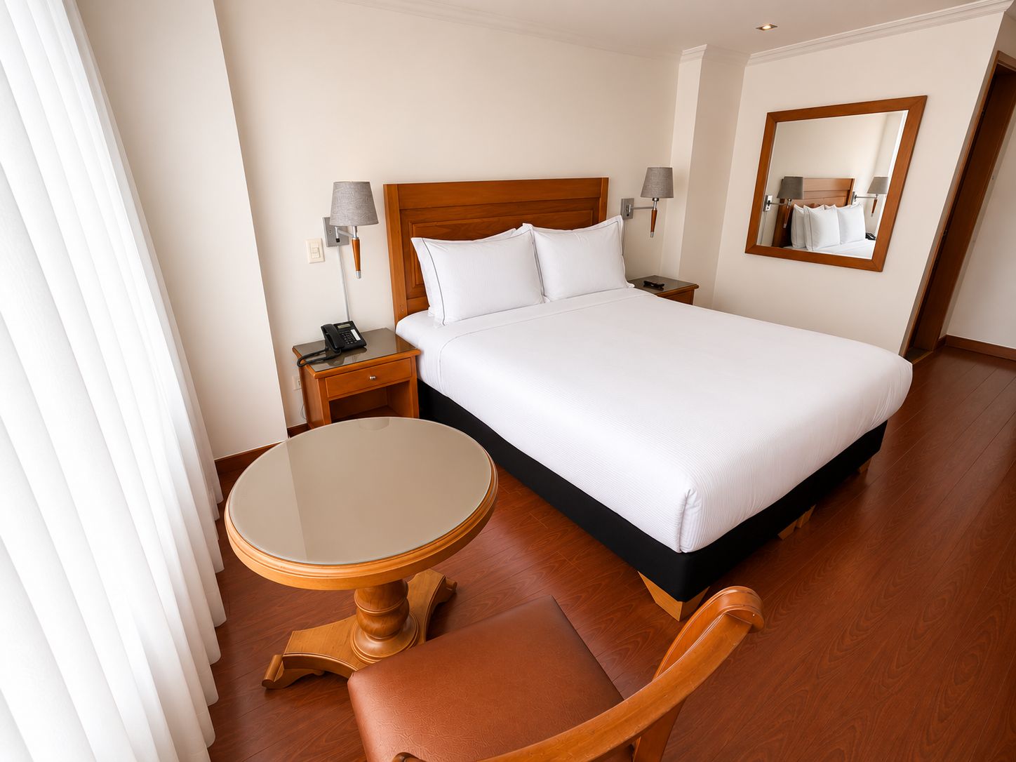 A hotel room with a bed, bedside tables, a round table, a chair, and a mirror.