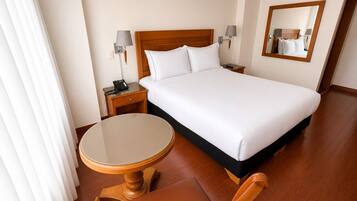 Una habitación de hotel con una cama, mesitas de noche, una mesa redonda, una silla y un espejo.