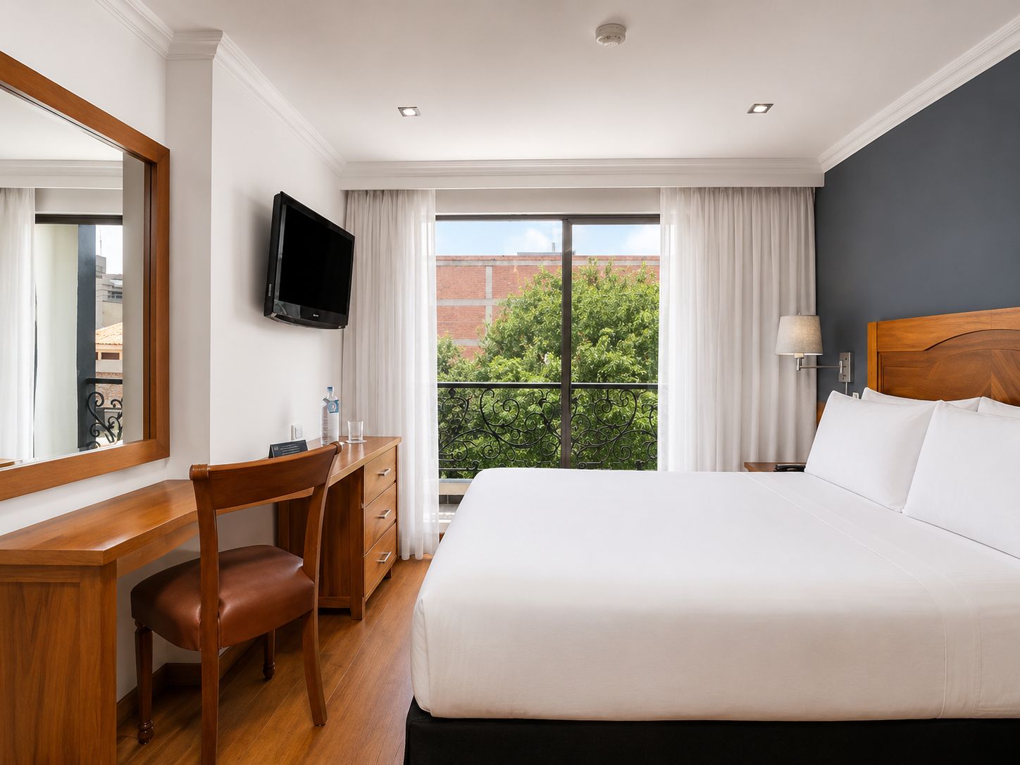 Habitación de hotel con cama, escritorio, silla, televisión y ventana con vistas a zonas verdes y edificios.