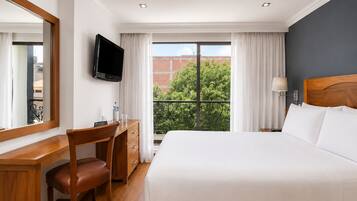 Habitación de hotel con cama, escritorio, silla, televisión y ventana con vistas a zonas verdes y edificios.