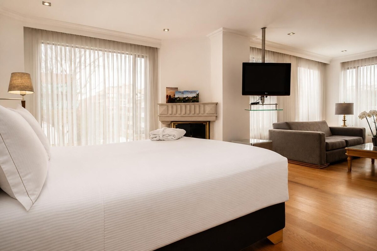 Habitación de hotel con una cama grande, televisión, chimenea, zona de estar con sofá y sillones, y una mesa de centro.