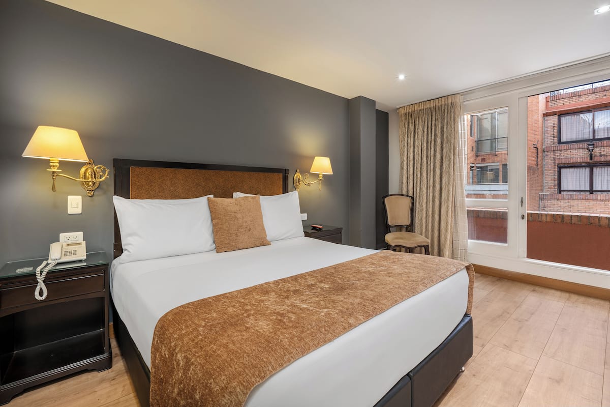 A hotel room with a large bed, two bedside lamps, a telephone, and a view of a brick building through a window.