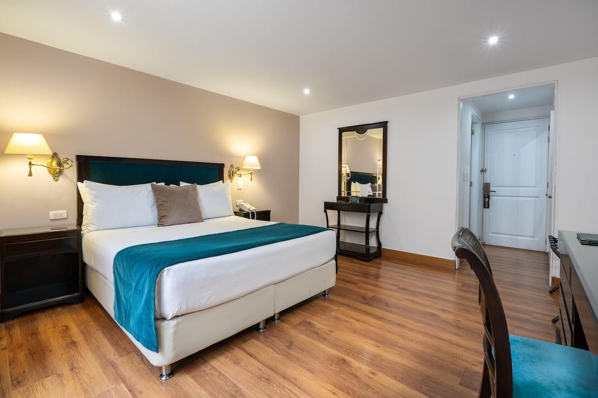 A hotel room with a bed, bedside lamps, a desk, a chair, a mirror, and a door.