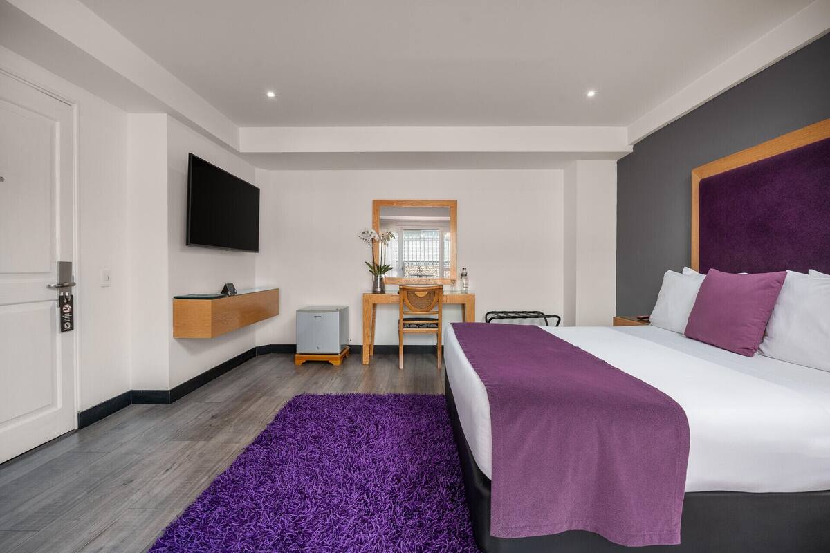 A modern hotel room with a bed, a desk, a chair, a TV, and a small plant.