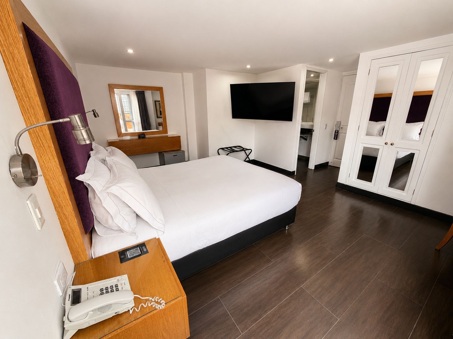 Habitación de hotel con cama, teléfono en la mesita de noche, espejo, televisor montado en la pared y armario con puertas de vidrio.