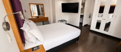 Habitación de hotel con cama, teléfono en la mesita de noche, espejo, televisor montado en la pared y armario con puertas de vidrio.