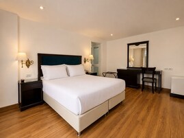 Una habitación de hotel con cama, mesitas de noche, escritorio, silla, espejo y radiador.