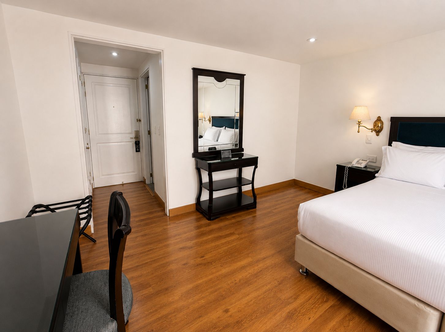 A hotel room with a bed, a desk, a chair, a mirror, a nightstand, and a lamp.