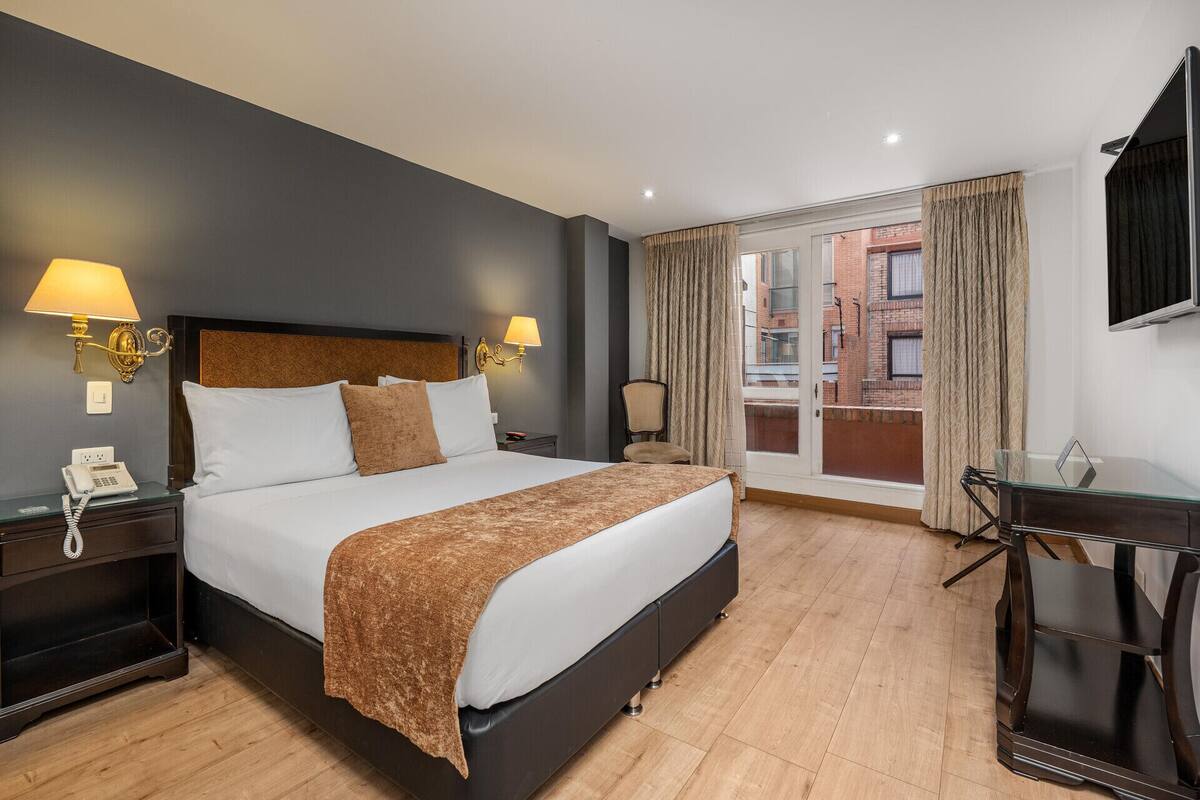 A hotel room with a bed, bedside lamps, a telephone, a chair, a desk, and a view of the outside.