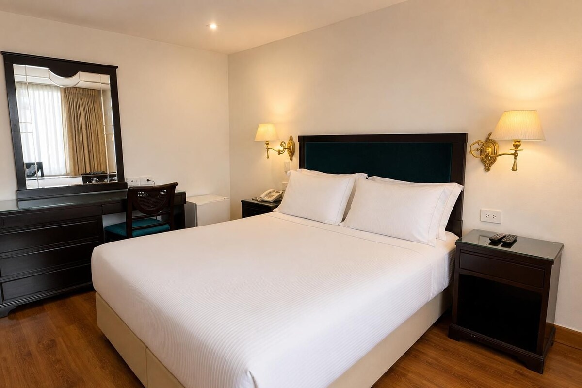 Habitación de hotel con una cama grande, cómoda, un tocador, un espejo, dos lámparas de noche y una mesita de noche con teléfono y controles remotos.