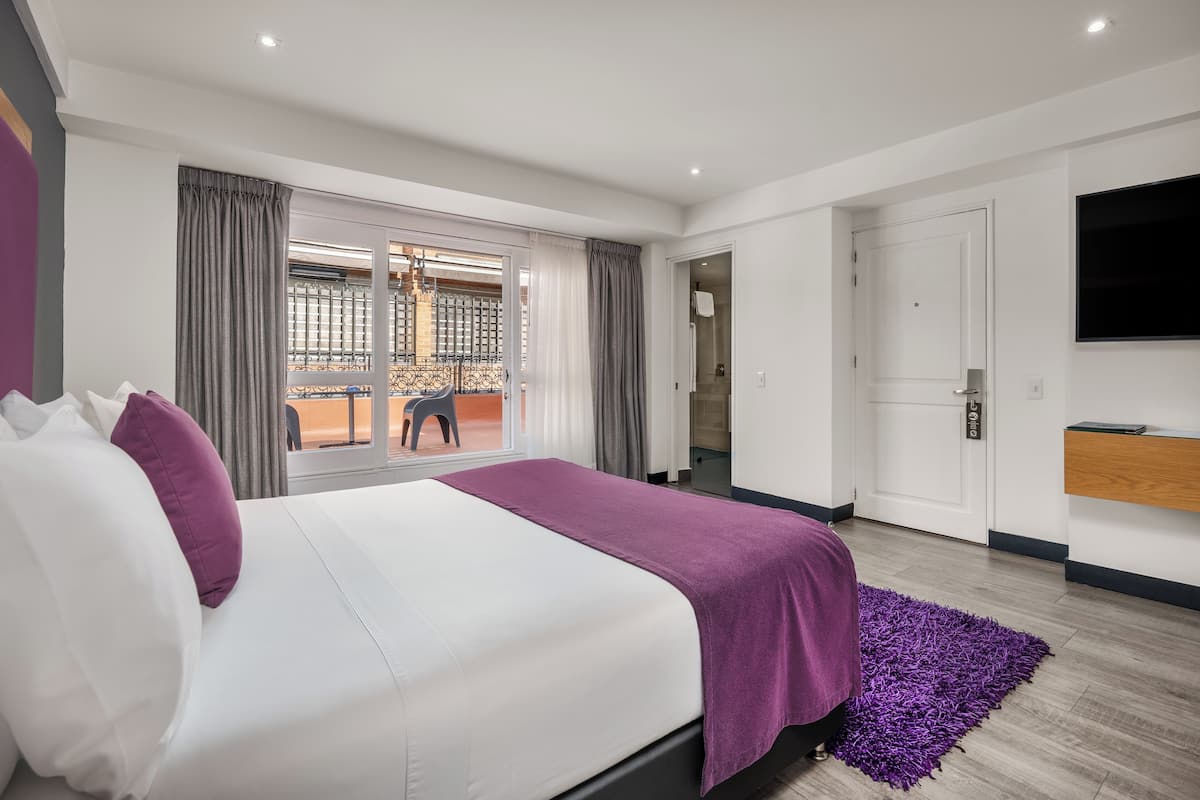 A modern hotel room with a large bed, a balcony with a view, a TV, and a wooden floor.