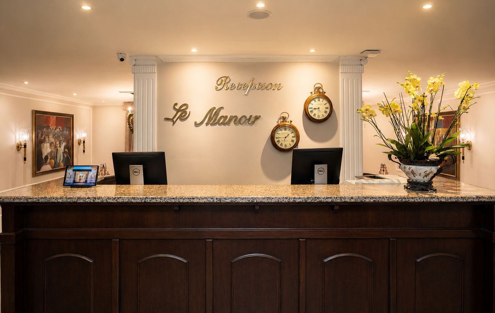 Zona de recepción de un hotel con mostrador de mármol, dos monitores de computadora, un reloj y una planta en maceta.