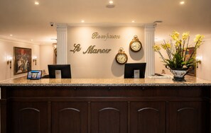 Zona de recepción de un hotel con mostrador de mármol, dos monitores de computadora, un reloj y una planta en maceta.