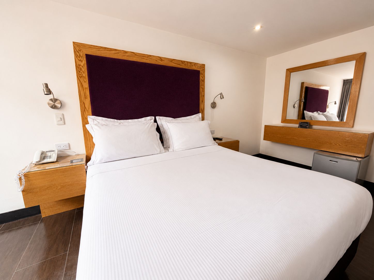 Quarto de hotel com uma cama grande, mesa de cabeceira com telefone, criadeira, espelho e pequena escrivaninha com luminária.