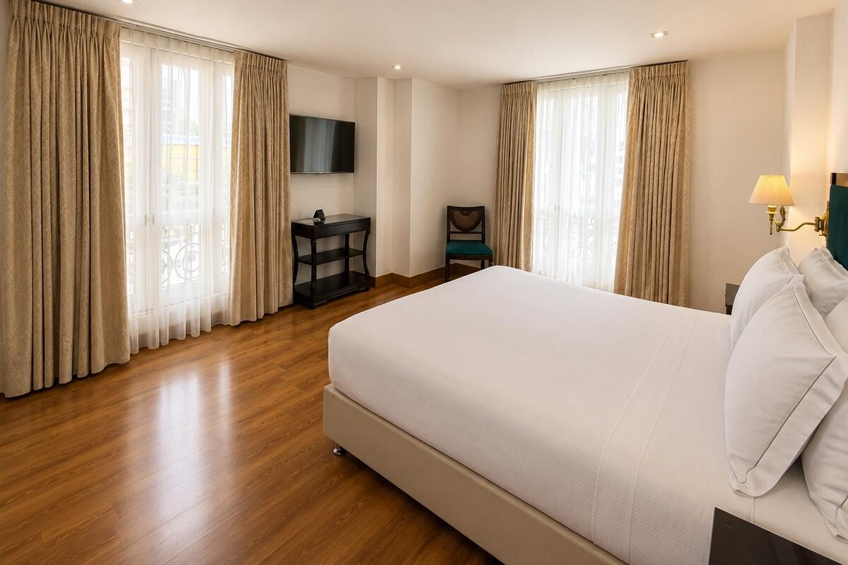 A hotel room with a large bed, wooden flooring, a TV on the wall, a small table, and curtains.