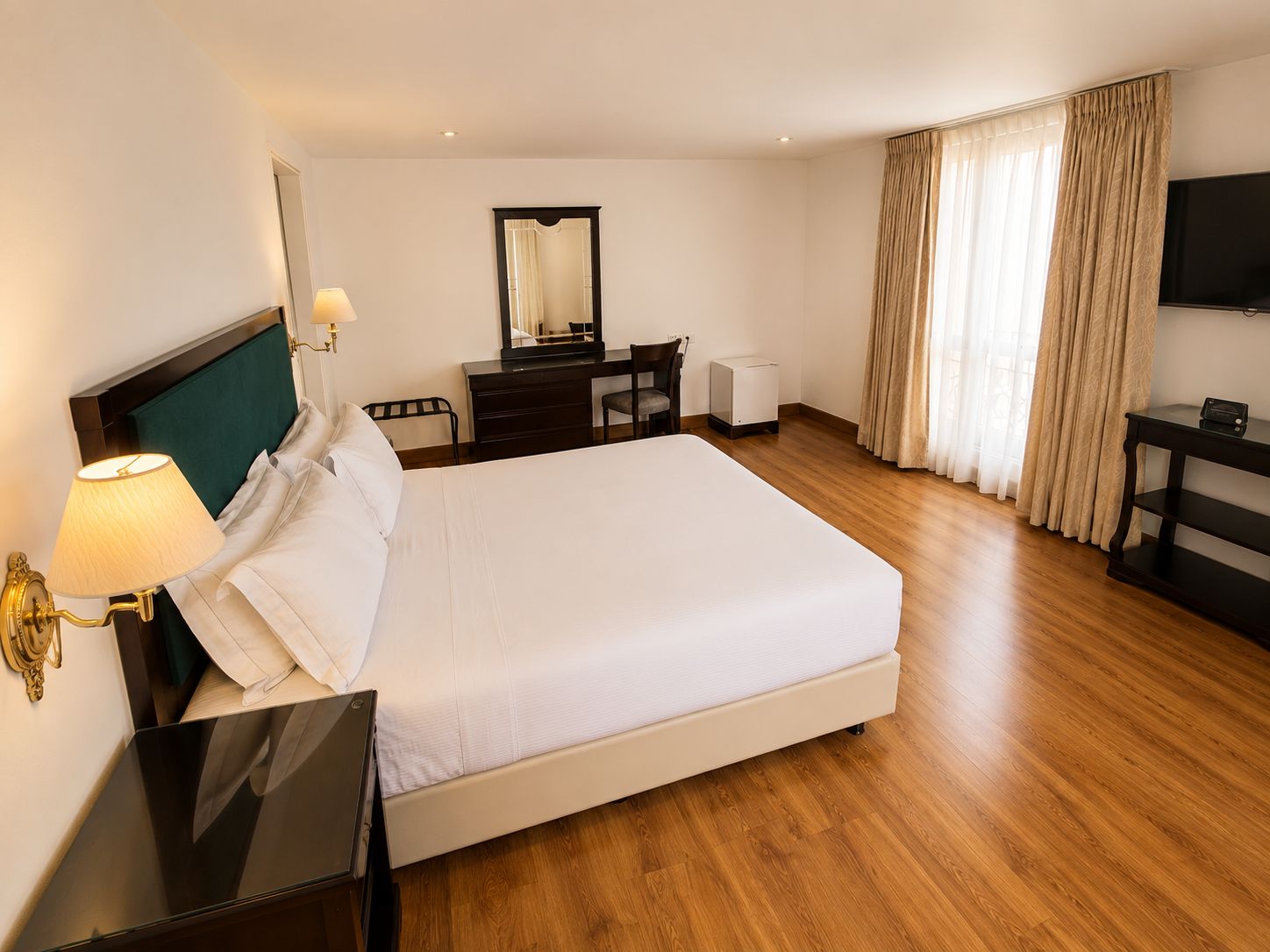 A hotel room with a bed, bedside table, lamp, desk, chair, mirror, television, and curtains.