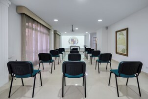 Un salón de conferencias con filas de sillas, una pantalla de proyector y un cuadro en la pared.