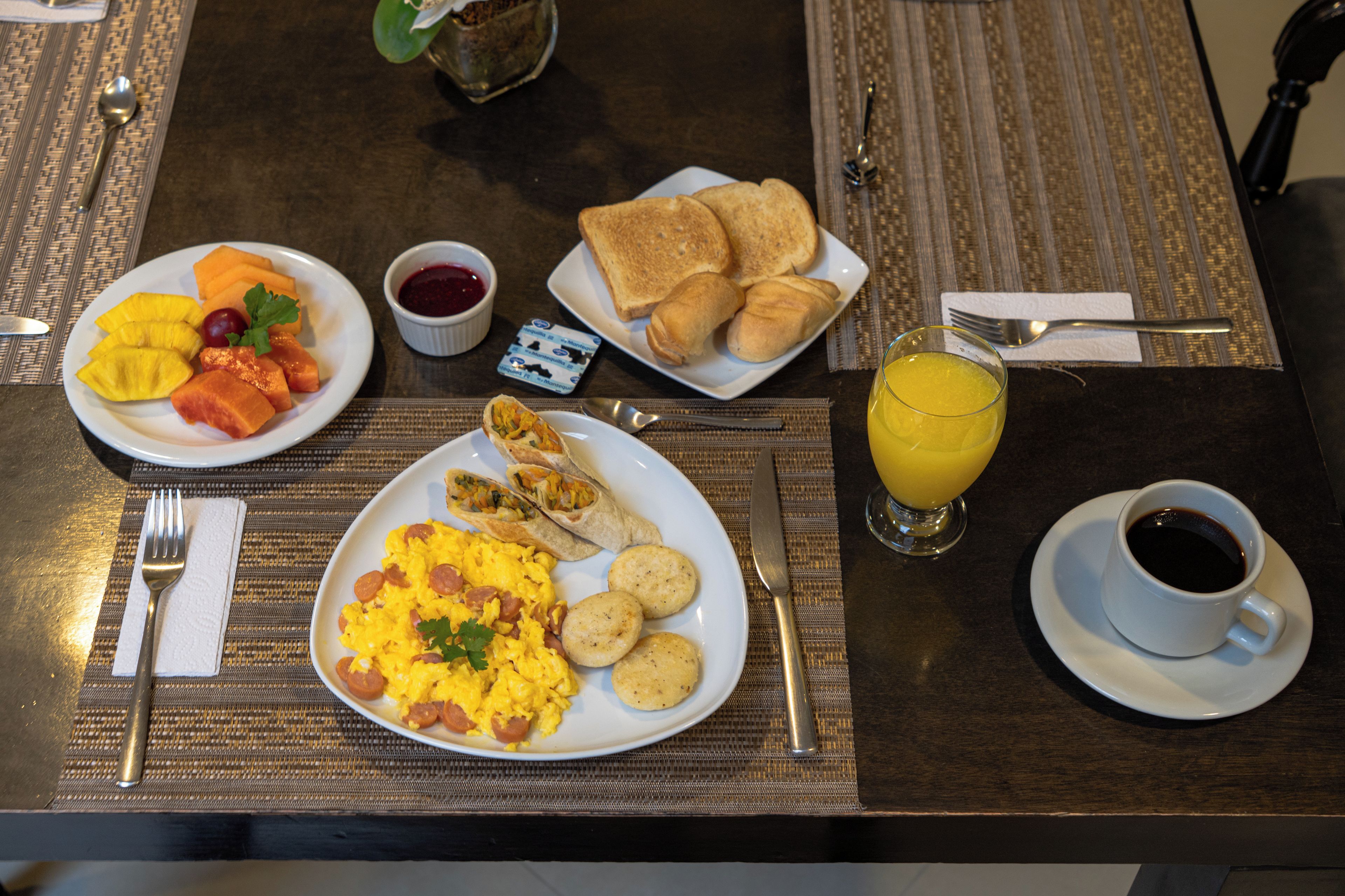 Een ontbijttafel met roerei, toast, fruit, een glas sinaasappelsap en een kop koffie.