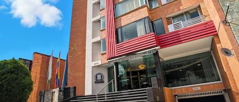 Un hôtel à plusieurs étages, doté d’une façade rouge et blanche bien visible, d’un escalier menant à l’entrée et, en arrière-plan, d’un ciel bleu clair.