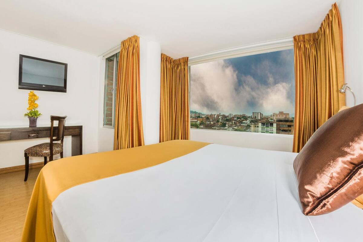 Habitación de hotel con cama, escritorio, silla, televisor y vista a la ciudad.