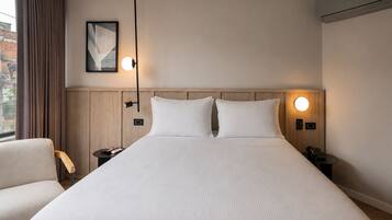 Habitación de hotel con una cama grande, dos mesitas de noche, una silla, un cuadro en la pared y una ventana con cortinas.